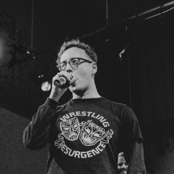 A picture of a white man with glasses in a jumpers that says "wrestling resurgence" speaking into a handheld microphone.