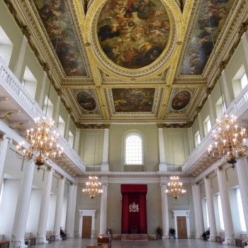 A large room with a painted ceiling with golden frames around the detailed pictures. Two galleries on teh right and left, the top having windows in and the bottom with columns coming down. Four chandeliers come down from the ceiling and the far end of the picture shows a high window with light coming through and below it a wooden door.