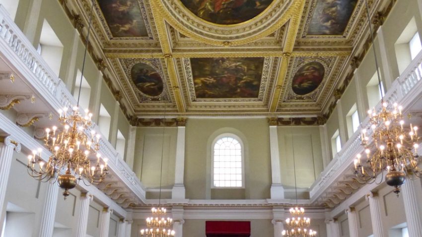 A large room with a painted ceiling with golden frames around the detailed pictures. Two galleries on teh right and left, the top having windows in and the bottom with columns coming down. Four chandeliers come down from the ceiling and the far end of the picture shows a high window with light coming through and below it a wooden door.