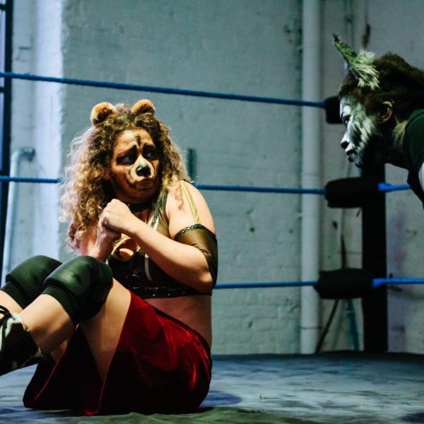 A wrestler with their face painted like a bear crouches in fear, wearing shin-length boots and kneepads and red shorts, away from the profile of a figure on their haunches with their face painted blue and grey and with ears on their head. In the background is the corner of the boxing ring and a large window.