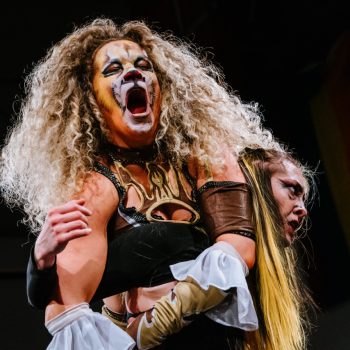 A woman with long curly hair with her face painted like a lion holds another person with long yellow and black hair under her left arm in a headlock. The person with the lion-painted face wears frilly cuffs and a wrestling outfit with gold flames and no arms.