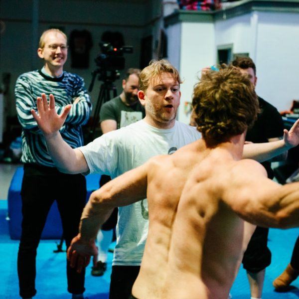 A white man with tousled hair holds both his hands up, wearing a white t-shirt. Another white man with their back to the camera moves as if to high-five one of them. In teh background, several people with their arms cross look on.