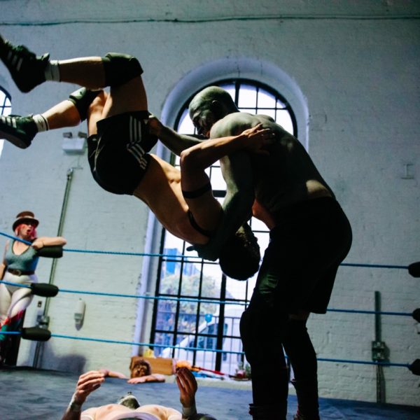 A large figure with their body and face painted holds another person mid-air, wearing just shorts and ankle-length boots, slamming them towards the floor of a boxing ring, on which the prostrate form of another wrestler is just about visible. Around the edges of the ring, other wrestlers, one with a painted face like a bear, look on. A woman in a bowler hat and skin-tight outfit with flared trousers looks on shouting.