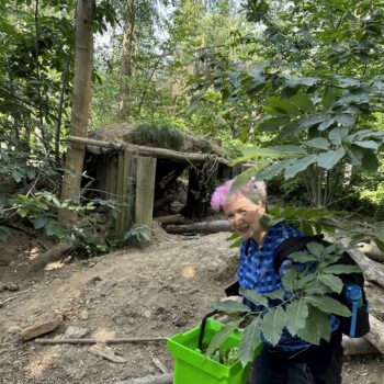 A woman with pink hair in a blue check shirt holds a green basket full of food. In the background is a makeshift set out of wood and dirt with greenery surrounding.